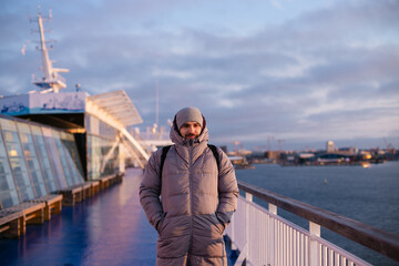 Handsome young adult man standing on a sun deck of a cruise ship, wearing warm grey puffer coat and a hat, Helsinki, Finland, Europe