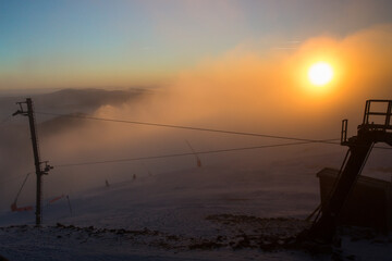 The sun rises over a snowy mountain landscape, casting warm golden hues across the foggy atmosphere.