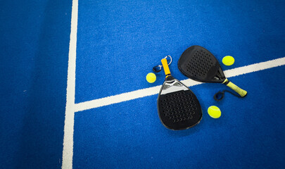 A padel or paddle tennis racket and a bright yellow tennis ball lie on a blue court. The net and a blurred figure of a player are visible in the background.