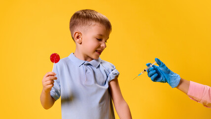 Boy holding red lollipop, smiling nervously while looking at syringe held by gloved hand against...