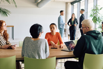 A diverse team of business professionals engaged in a discussion around a conference table in an office, while their colleagues collaborate in the background