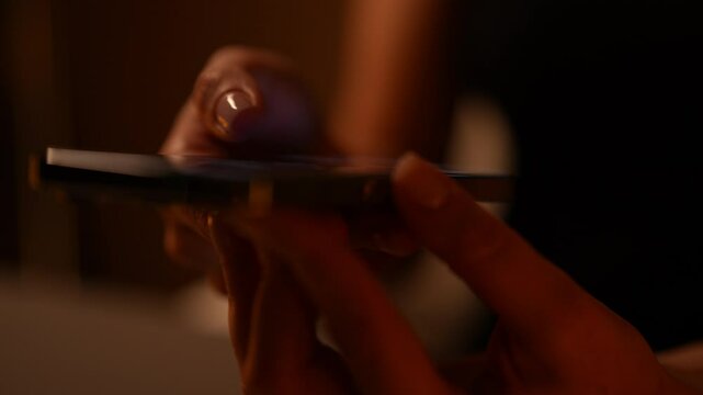 Close-up hands of unrecognizable woman scrolling and tapping on smartphone screen in dimly lit room. Female late-night mobile phone usage and engagement with online content. Shooting in slow motion.