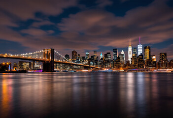 Fototapeta premium A night skyline of New York City with the iconic Brooklyn Bridge and the 911 memorial lights shining into the sky, reflecting on the calm waters of the East River