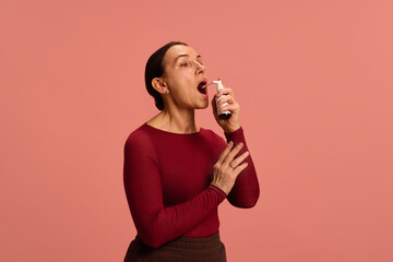 Mature woman in red shirt sprays throat medicine into her mouth with pained expression against pink...