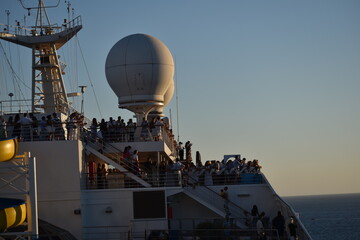 Crowd of people on a cruise ship deck at sunset with clear skies and calm ocean in the background The passengers enjoy the view as the ship sails through peaceful waters
