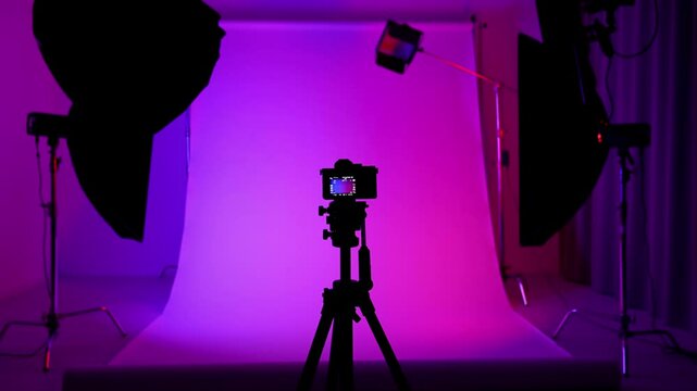 Professional studio for photo video production with modern lighting equipment, cyclorama background lighted with colorful neon light, man turns on camera on tripod.