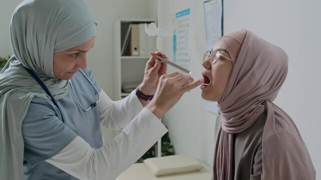 Female doctor in hijab performing oral examination with tongue depressor and penlight on Muslim patient in clinic