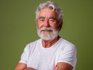 Portrait of a senior man smiling warmly against a plain green background, wearing a white t-shirt with arms crossed.
