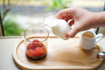 A woman's hand pours milk into a glass filled with strawberry ice cubes and espresso to make a strawberry latte.