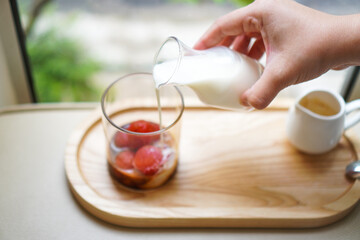 A woman's hand pours milk into a glass filled with strawberry ice cubes and espresso to make a strawberry latte.