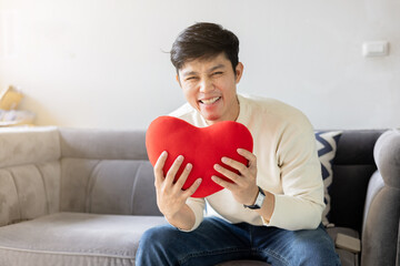 close up young asian man smile with happiness feeling and hold red color of heart shape pillow...