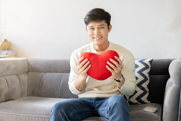 Fototapeta premium close up young asian man smile with happiness feeling and show red color of heart shape pillow while sit on couch in living room for good health and live better and valentine day and people lifestyle