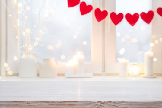 Bleached wooden table top in front of blurred window sill decorated with red heart garland for valentine day celebration