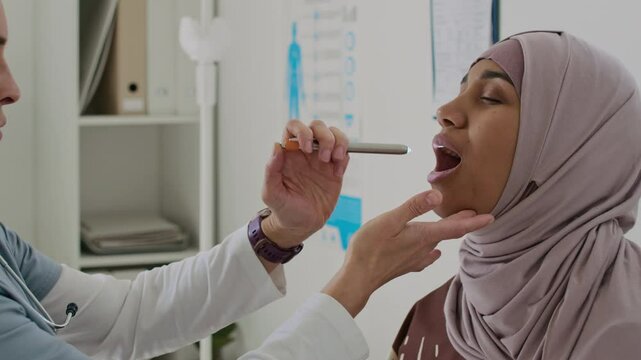 Female Doctor in Hijab performing throat check-up with special penlight, while her patient sitting on examination couch with her eyes closed