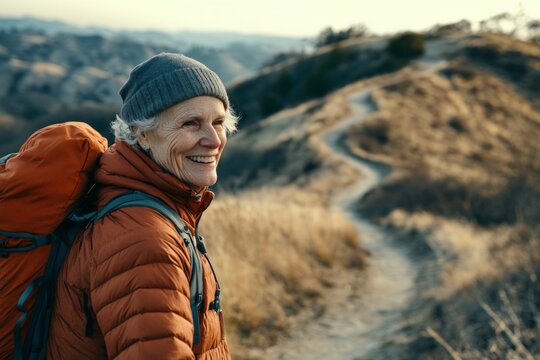An elderly woman in a hiking outfit and backpack smiles as she hikes on a scenic mountain trail, surrounded by dry grass and rolling hills on a clear day.