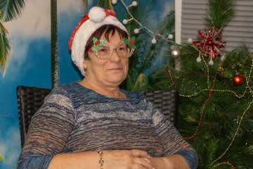 A smiling woman wearing a Santa hat and decorative glasses with holly accents sits near a Christmas...