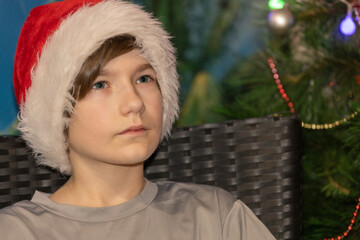 A young boy wearing a Santa hat sits in a wicker chair, gazing thoughtfully near a decorated Christmas tree with festive ornaments and lights.