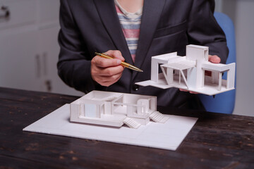 A businessman sits at a table at night, holding a model of a two-story white house. He reviews...
