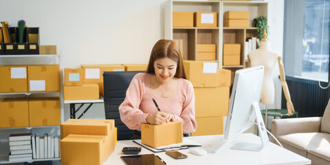 An Asian businesswoman sits at her desk, working on online sales and e-commerce marketing strategies. As a small business owner, she prepares and packages product boxes to ship to customers.