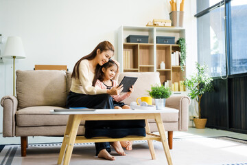 An Asian mother and her adorable daughter sit on the sofa in their living room, enjoying online learning on a tablet and reading books. A joyful family moment focused on education