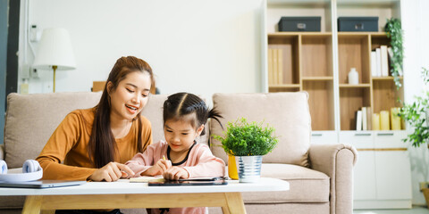 An Asian mother and her adorable daughter sit on the sofa in their living room, enjoying online learning on a tablet and reading books. A joyful family moment focused on education