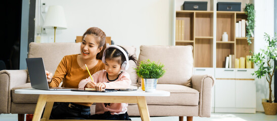 An Asian mother and her adorable daughter sit on the sofa in their living room, enjoying online learning on a tablet and reading books. A joyful family moment focused on education