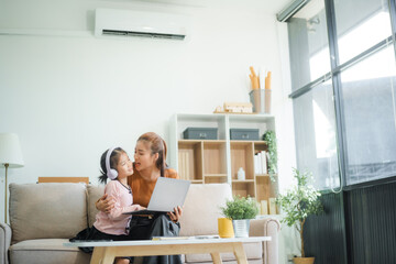 An Asian mother and her adorable daughter sit on the sofa in their living room, enjoying online learning on a tablet and reading books. A joyful family moment focused on education