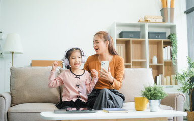 An Asian mother and her adorable daughter sit on the sofa in their living room, enjoying online learning on a tablet and reading books. A joyful family moment focused on education