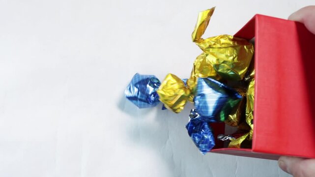 Male hands open yellow lid with bow of gift box full of gift candies wrapped in foil of different colors and pour candies on white surface, top view.