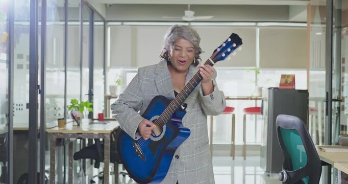 Indian Asian happy senior gen x business woman play guitar singing song indoor job place excited cute old silly crazy lady staff worker dancing enjoy day break time have fun joyful joy work office