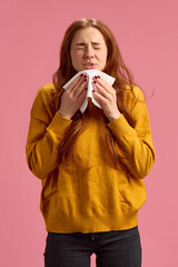 Young redhead woman in yellow sweater sneezing on pink studio background. Cold and flu symptoms, allergy, sinus infection, inflammation. Concept of illness, medicine, treatment, health care
