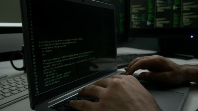 Close-up shot of unrecognizable programmer writing code on laptop, while sitting at desk, surrounded by monitors with information