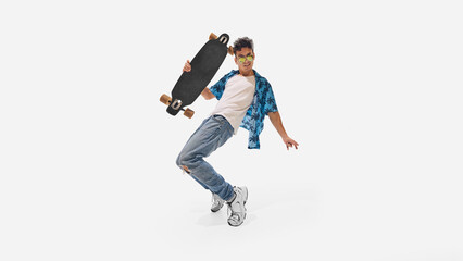 Skateboarder balancing, holding skateboard in one hand while leaning sideways in ripped jeans and blue shirt against white studio background. Concept of success, professional development, movement. Ad