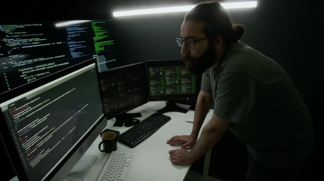 Long-haired, bearded man intensely studying four computer monitors displaying lines of code in dimly lit programmer workspace