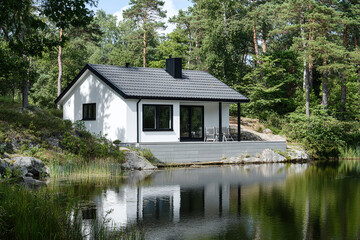 A simple, white house with a black roof and windows, situated in the middle of nature and surrounded by trees on one side, overlooking a body of water and a lake.The house is in the Scandinavian style