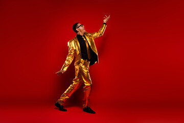 Young man in a golden suit and black shirt striking a dynamic pose against a vibrant red background.