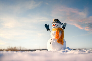 Smiley snowman in knitted hat and yellow scarf with hands up on snowy field. Warm sunset sky with...
