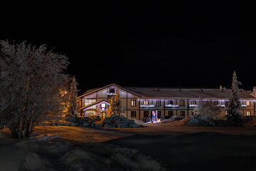 Snowy winter night scene with illuminated streetlights, frost-covered trees and a building on the side. Peaceful, cold and serene winter atmosphere. Cozy cottage in countryside
