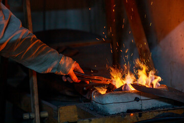 A blacksmith holds a billet in a pair of tongs over the stove