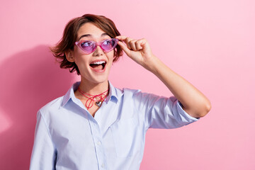 Portrait of astonished cheerful girl arm touch sunglass look empty space isolated on pink color...