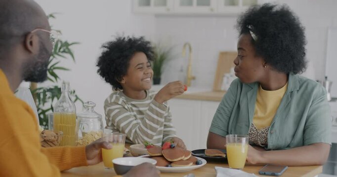 Loving son feeding mother with strawberry talking to mom enjoying breakfast at home with family. Childhood and motherhood concept.