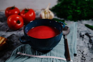 beetroot or tomato soup served in dark blue bowl, kitchen cloth on gray background, fresh tomatoes, garlic, roasted slice of bread