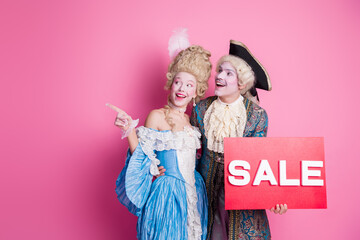 Cheerful couple in elegant vintage costumes poses with a sale sign against a vibrant pink backdrop