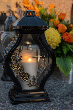 A beautiful candle shines on the grave, in the background yellow chrysanthemums and orange roses.