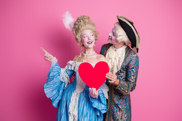 Young couple in elegant baroque costumes holding a heart against a pink background, embodying classic romance