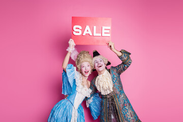 Joyful couple in extravagant costumes holding sale sign against vivid pink background, celebrating with laughter and fun at a themed party