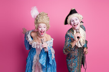 Lively couple in antique costumes with a humorous flair pose joyfully on a vibrant pink backdrop, embodying elegance and fun