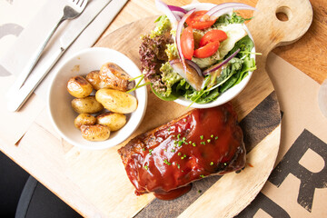 Pork ribs with barbecue sauce at an angle served on a cattail with cherry tomato salad and souté potatoes in the background top view