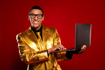 Charismatic young man in a golden suit holding a laptop against a vibrant red backdrop