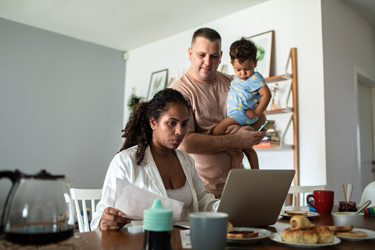 Stressed mother working on laptop while father entertains toddler during breakfast at home